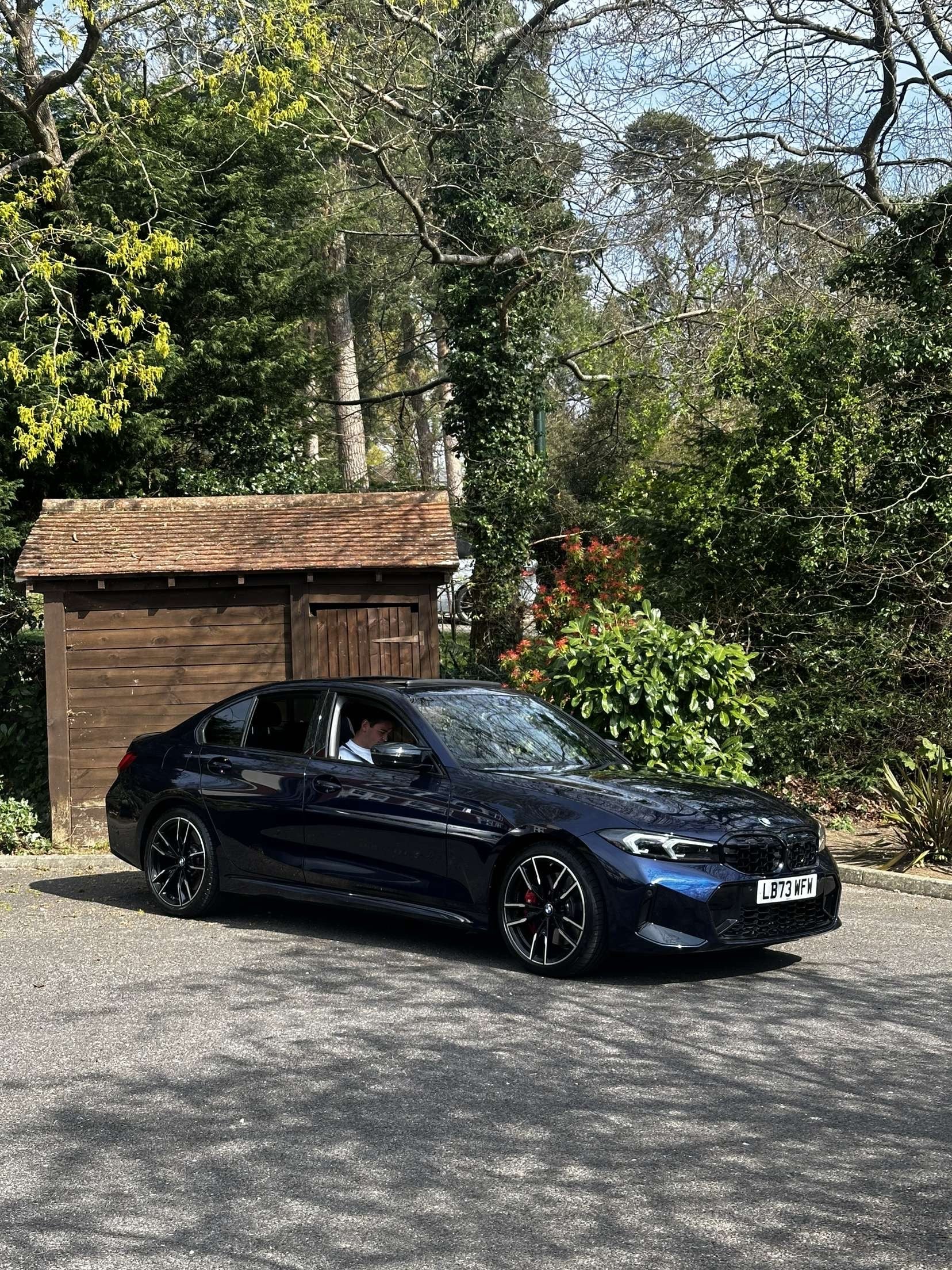Black car parked on a road with greenery and a wooden shed in the background